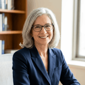 Synaptigen customer review profile photo of a smiling older woman with grey hair and glasses wearing a blue blazer.